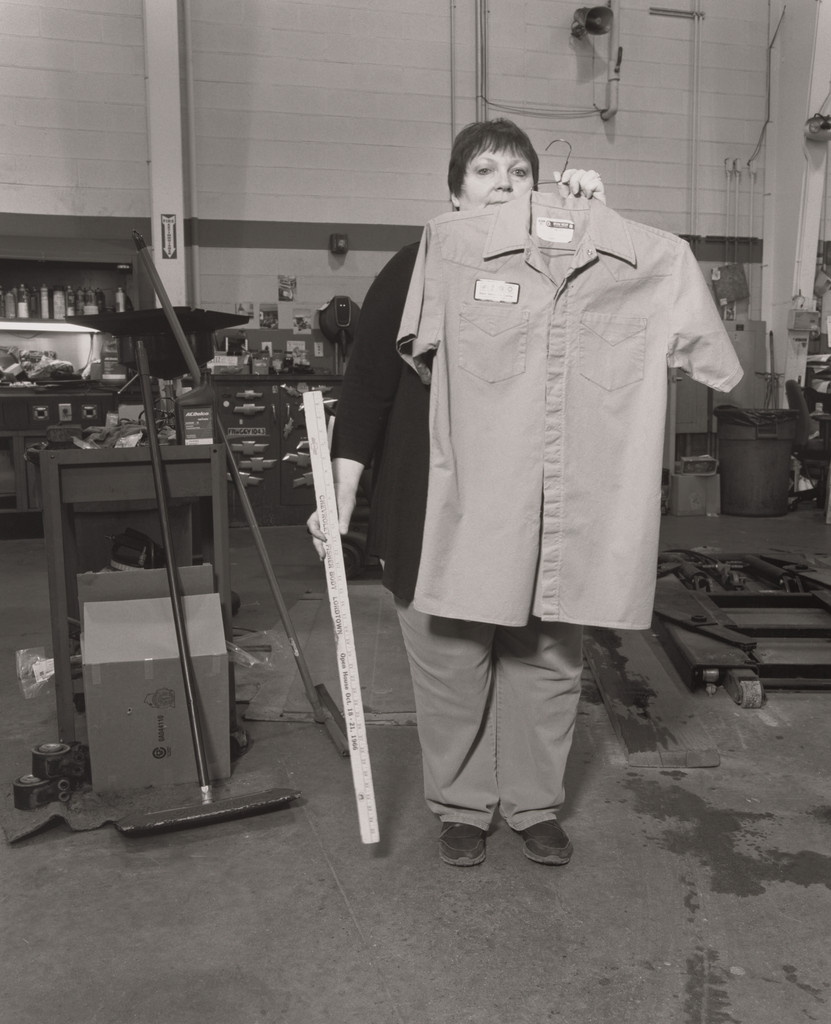 Lisa Cartwright, Service Cashier, holding her father’s work shirt and his Chevro-let Fisher Body antique ruler, inside the maintenance shop at Sweeney Chevro-let Buick GMC car dealership, Board-man, OH